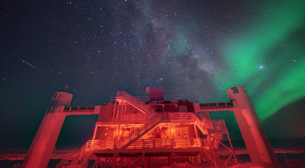 Laboratório do observatório IceCube à noite, na Antártida, com luzes na construção e céu estrelado.
